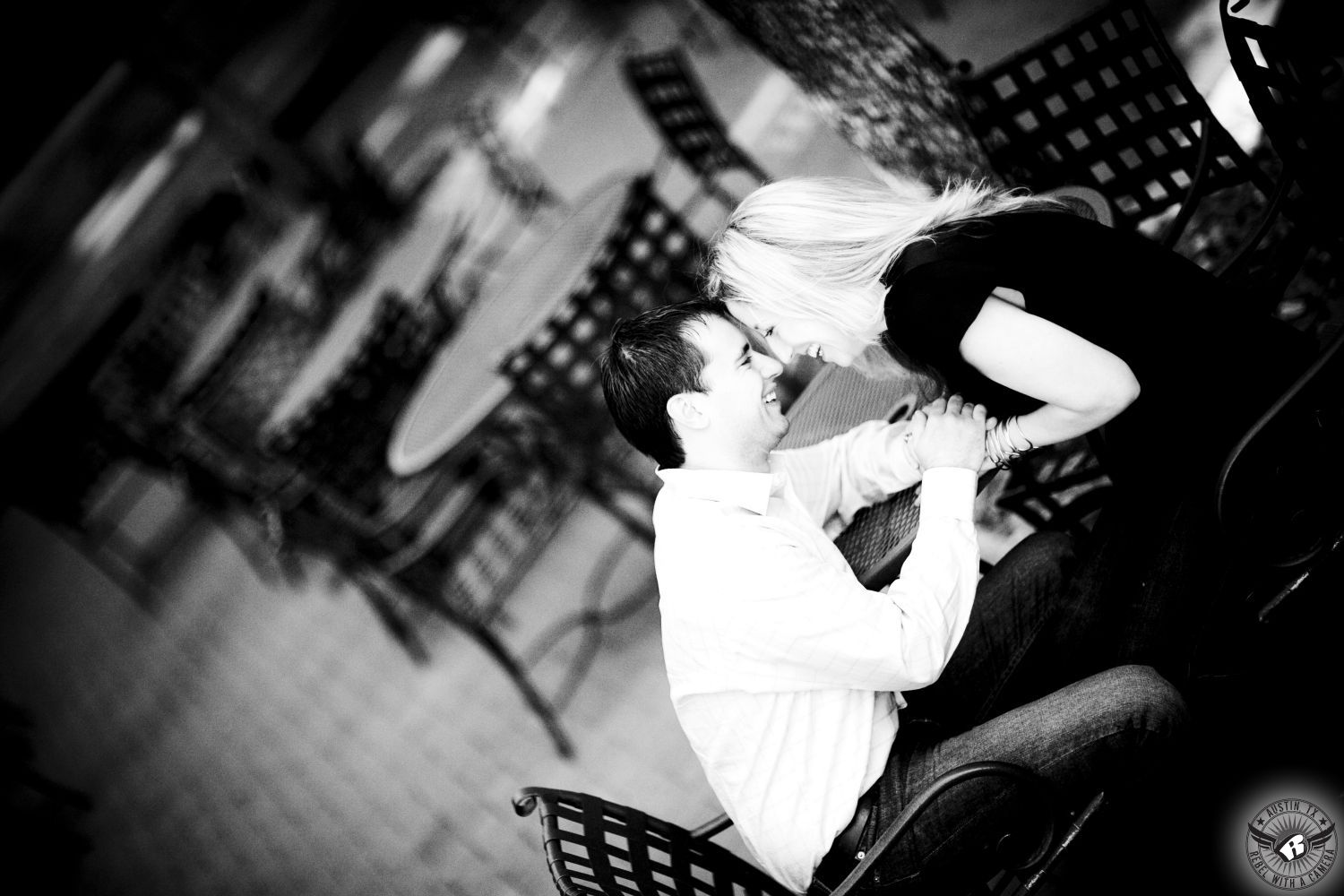 Blond girl wearing a black short sleeve blouse and black jeans laughs and holds the hands of a guy with dark hair wearing a white button up dress shirt and light jeans sitting at an outdoor  table in an open air bar on Third Street in this friendly engagement portrait in downtown Austin.  